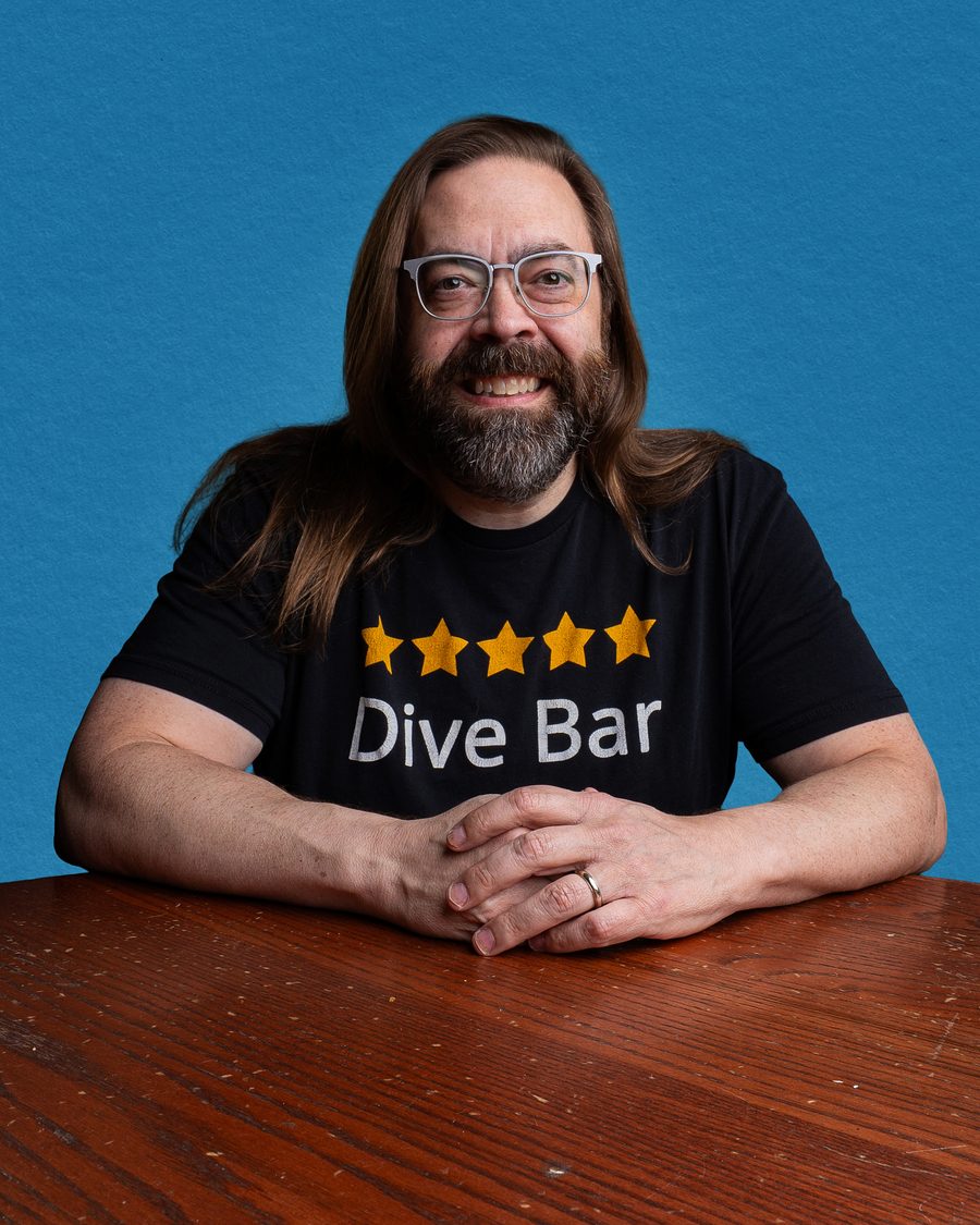 Gabriel Rutledge seated at a wooden table in a black five-star ‘Dive Bar’ t-shirt, hands folded, against a blue backdrop